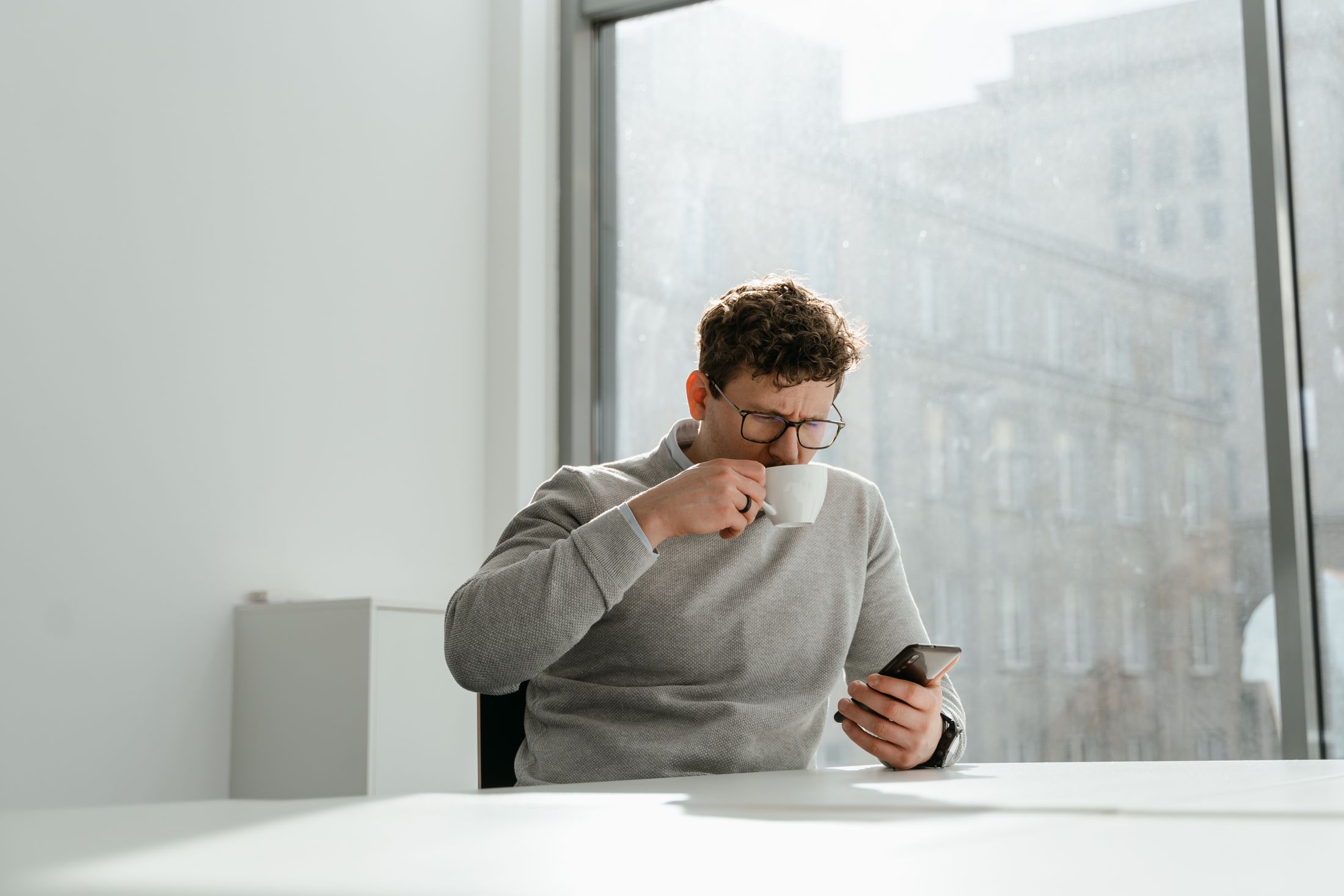 A man with a coffee checking his phone at a bright window